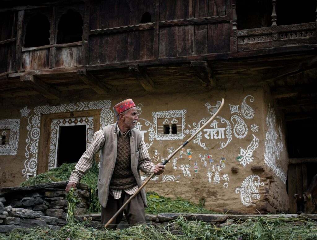 Elderly man in traditional attire in an Indian village setting, holding a stick.